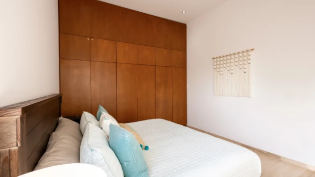 cama decoracion macrame dunas condo Bed with white linens, decorative cushions, and macrame wall hanging next to a large wooden closet.
