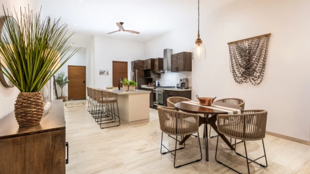 comedor cocina dunas condo Round dining table with woven chairs next to a dark fitted kitchen and decorative macrame.